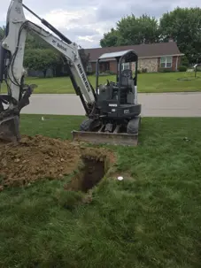 : A compact excavator preparing a clean water line entry pit on a green suburban lawn.