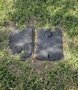 Two rectangular black plastic utility boxes sitting flush in a green grass lawn, commonly used to house irrigation valves or backflow preventers.