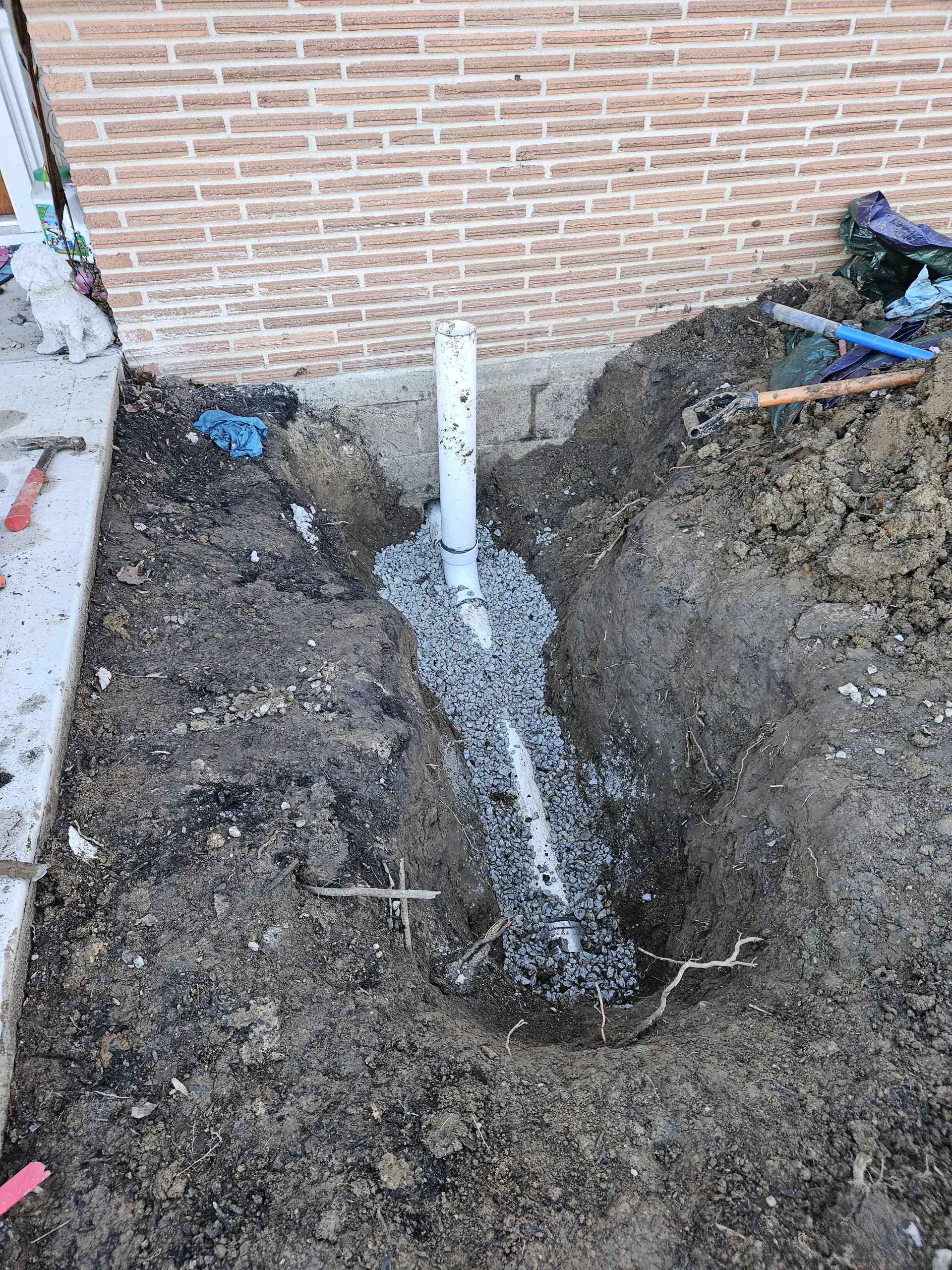 A trench excavated in a Dayton, Ohio yard showing a newly installed white PVC two-way exterior sewer cleanout pipe bedded in gravel.