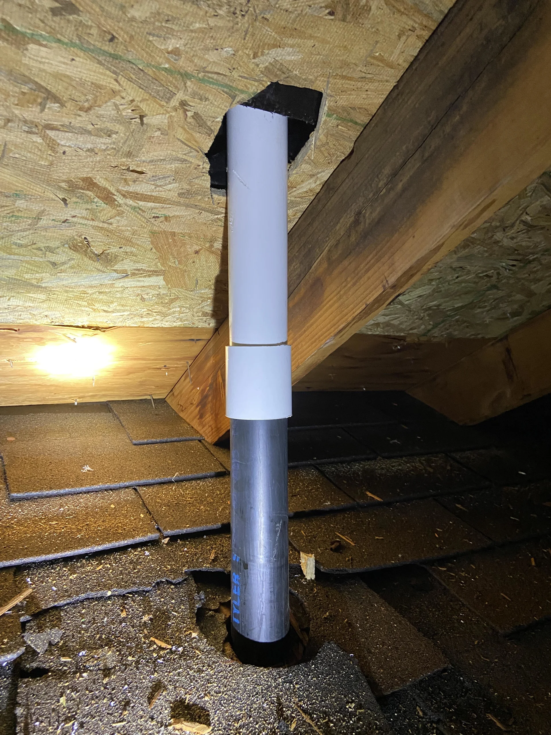 An interior view of a residential attic showing a PVC plumbing vent stack pipe passing through the wooden roof decking and shingles.