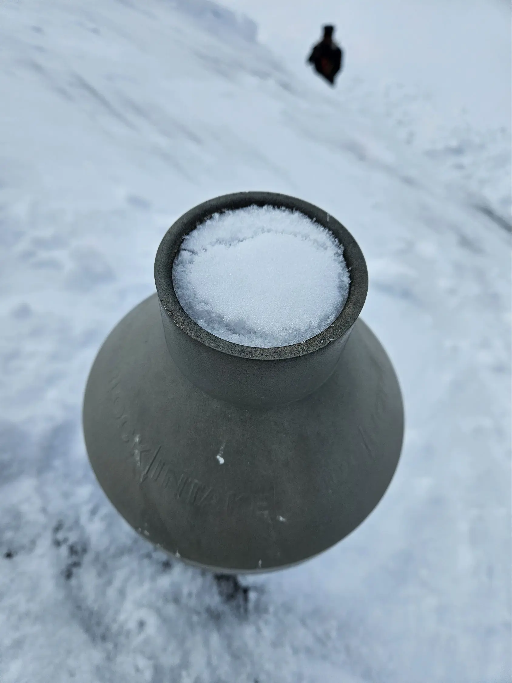 A metal roof vent pipe with a protective weather cap installed on a shingled residential roof against a bright blue sky.