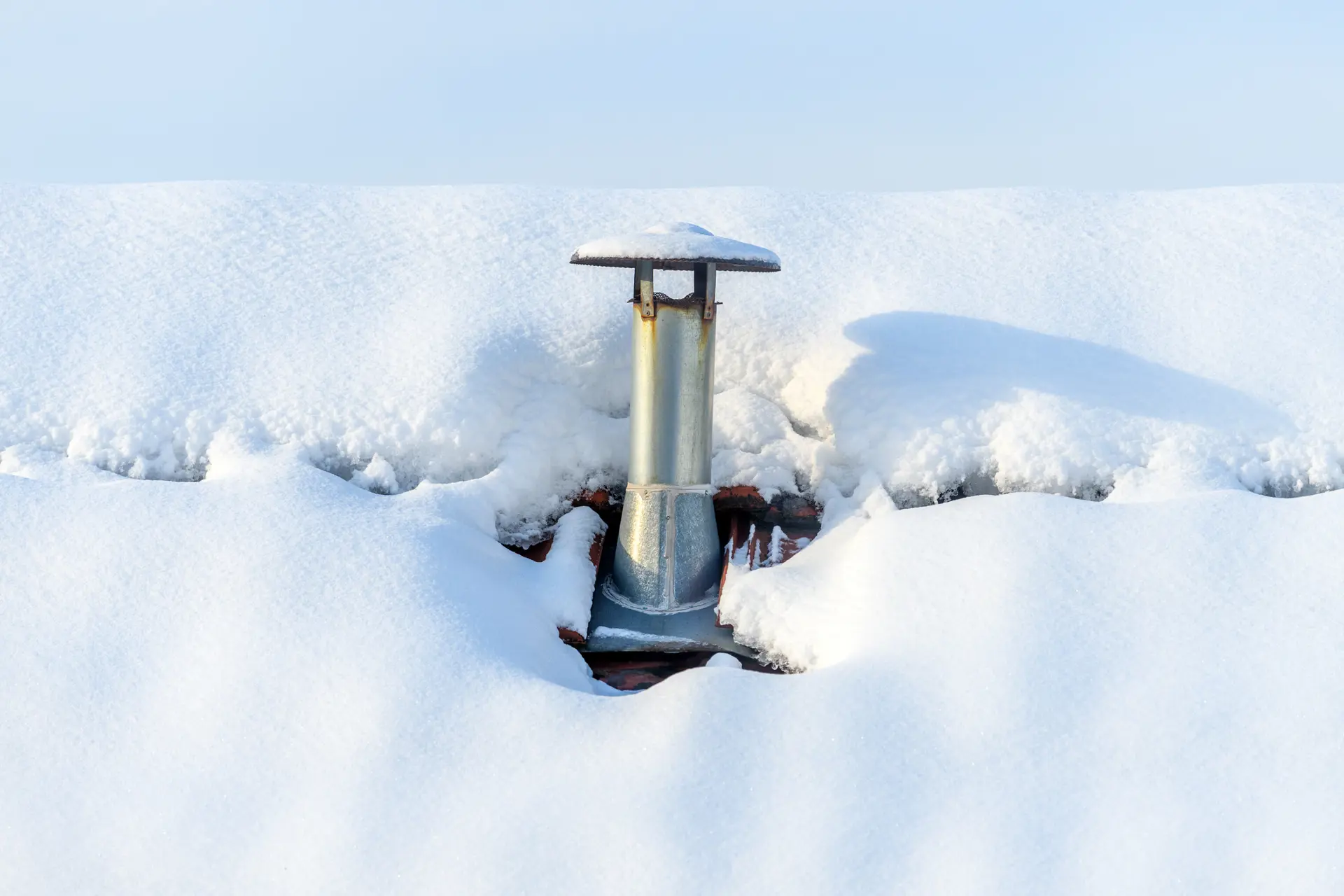 A metal roof vent pipe sticking out of a thick layer of heavy white snow on a residential roof during winter.