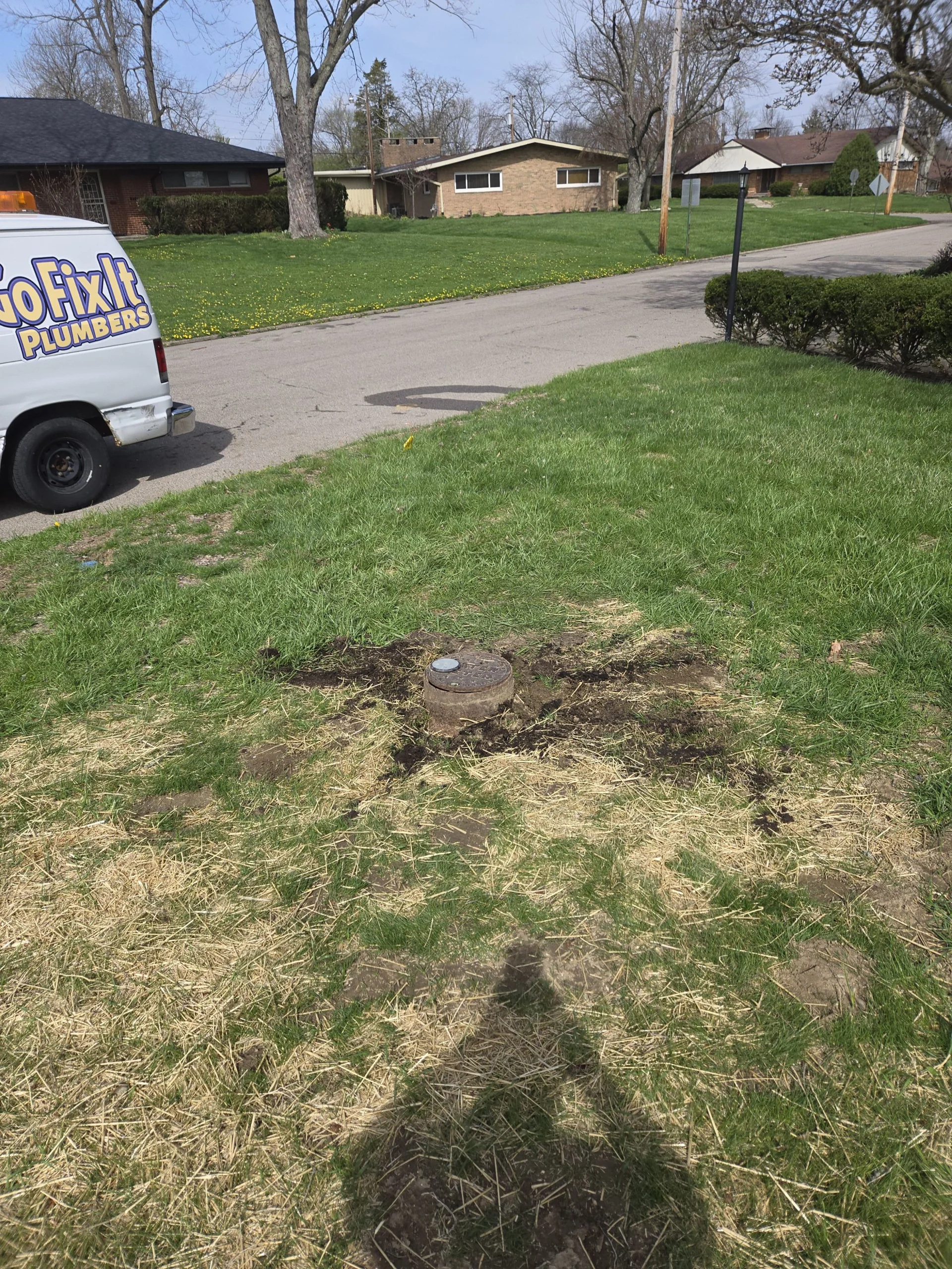 A round metal water meter cover sitting in a residential grass lawn near the street with a Honey Go Fix It plumbing van parked in the background.
